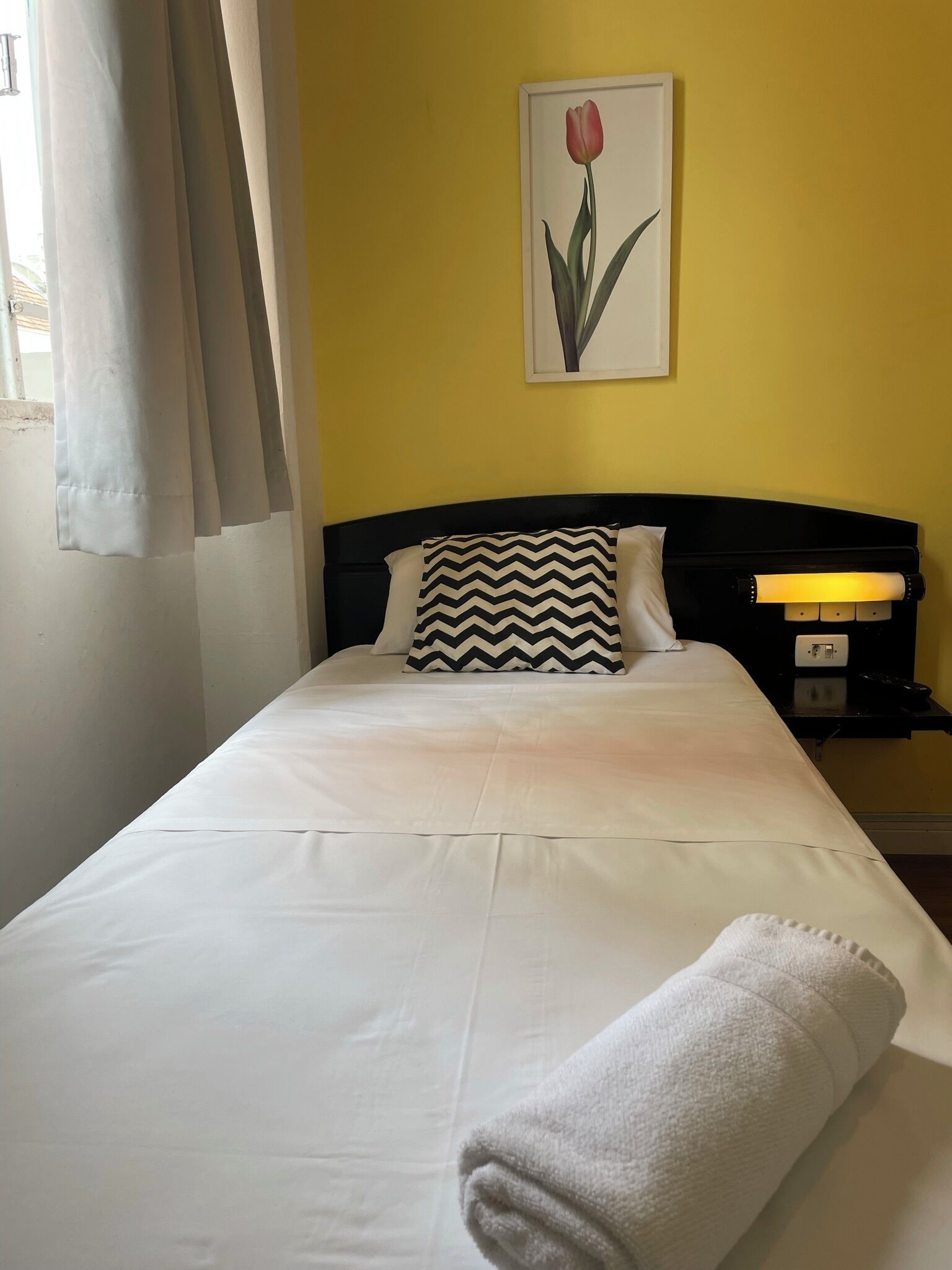 Cama bem arrumada com lençóis brancos, cabeceira preta e uma única almofada com padrão em zigue-zague. Uma toalha branca dobrada está no pé da cama.