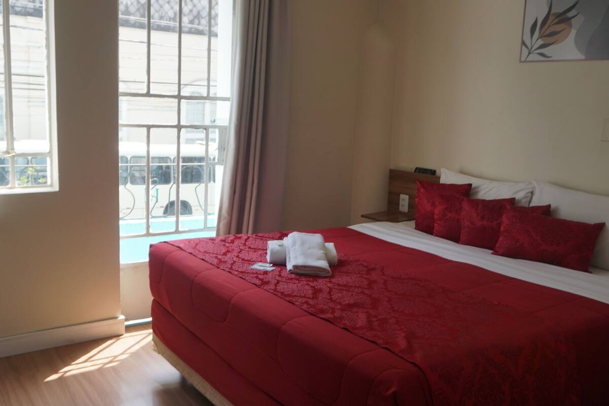 A hotel room with a large bed, red bedding, a nightstand, and a window with a view of a building and a bus.