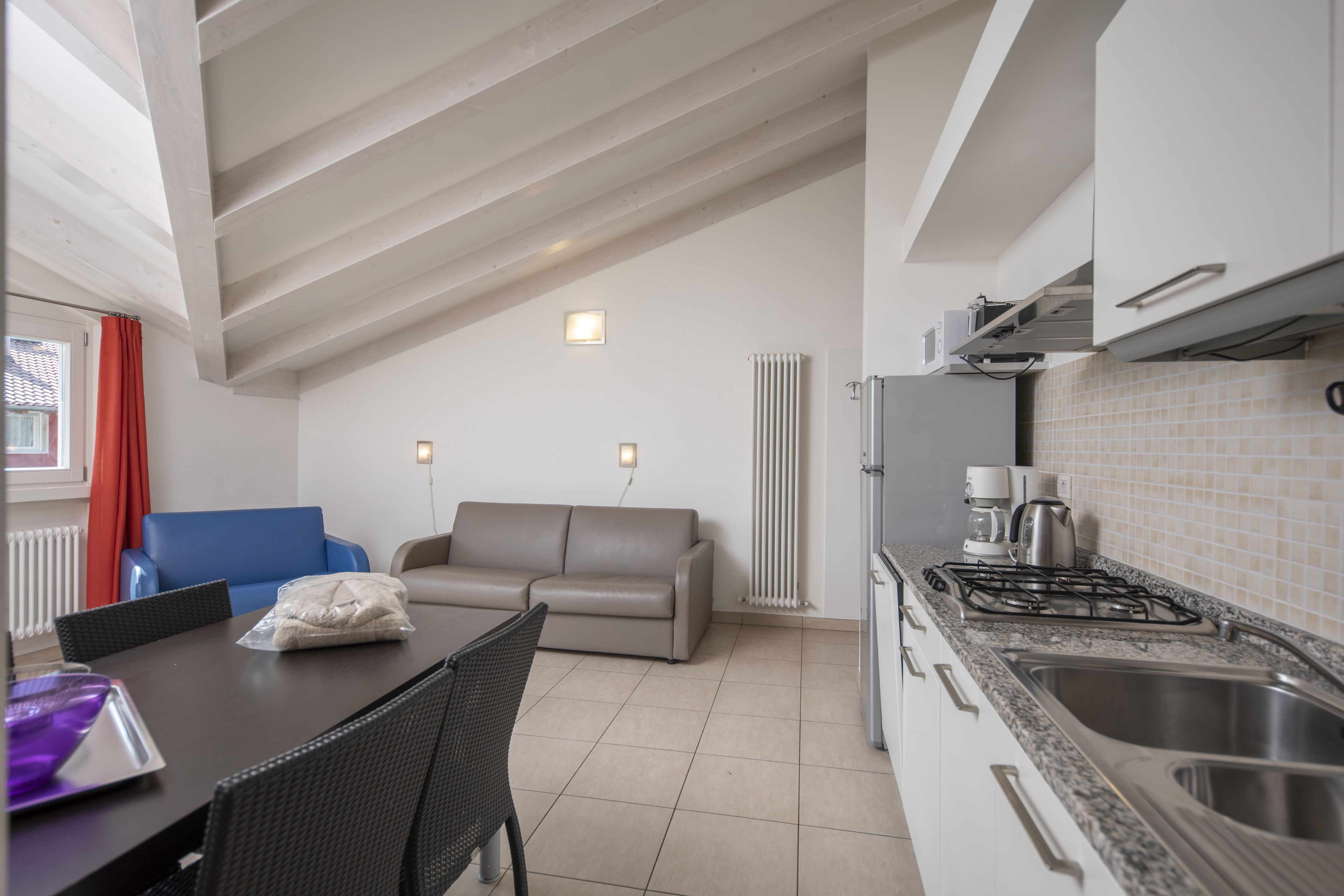 A modern kitchen with a gas stove, sink, and white cabinets. There is a dining area with a table and chairs, and a sofa in the background.