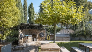A patio area with a stone barbecue station, wooden seating, and a tree providing shade.