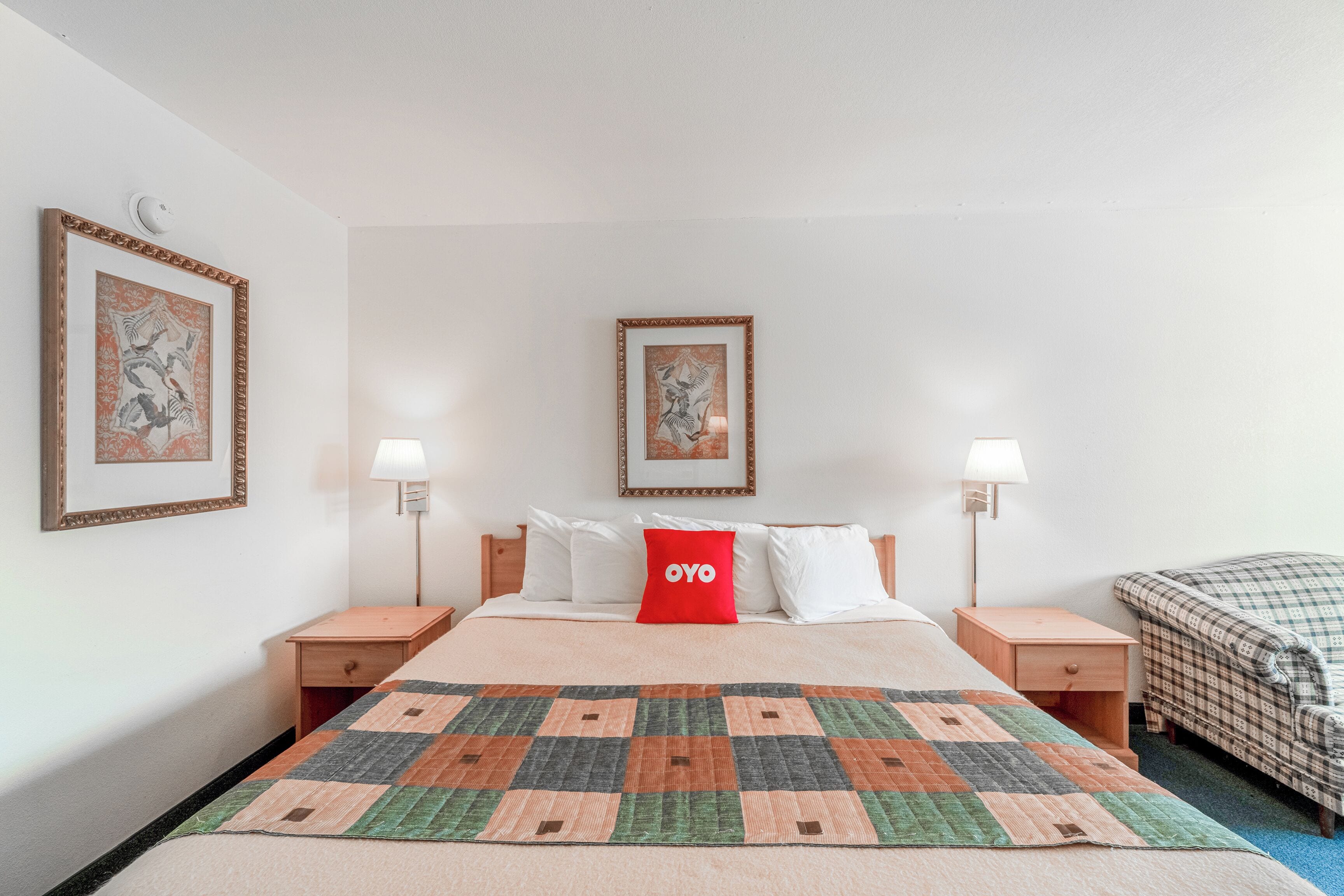 Habitación de hotel ordenada, con una cama grande, dos mesitas de noche, un cojín rojo y dos lámparas de pared.
