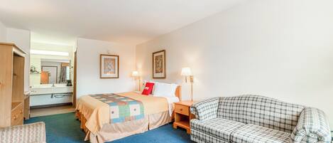 A hotel room with a bed, a sofa, a wooden wardrobe, a mirror, and a bedside table with a lamp.