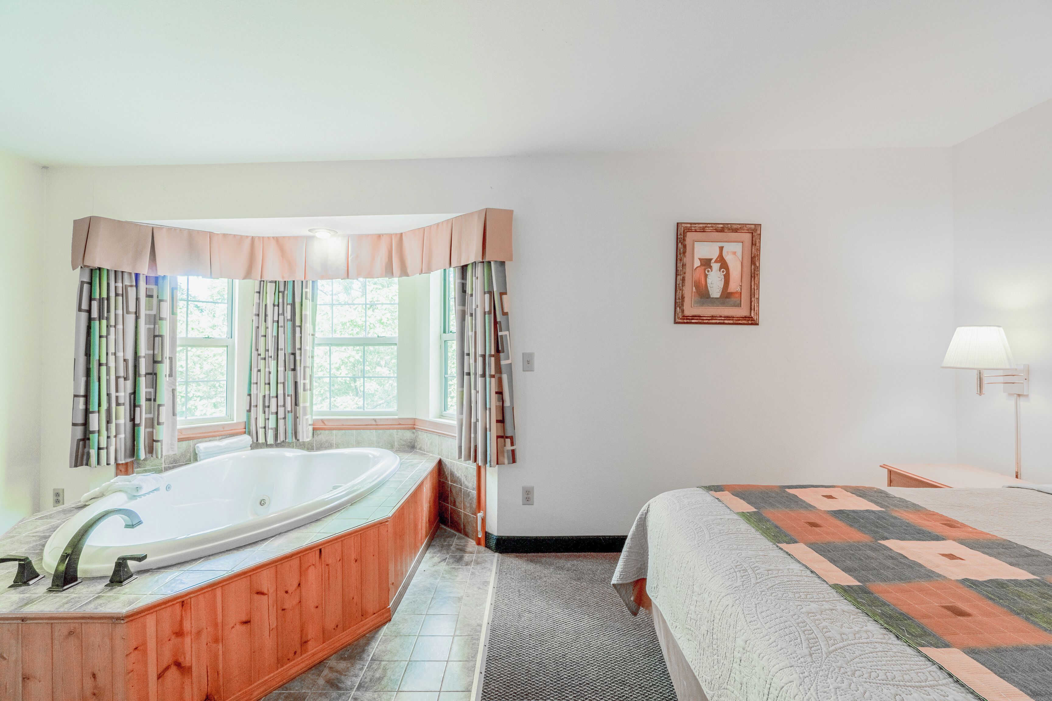 Un baño con una bañera independiente, un ventanal grande con cortinas, un tocador de madera y una cama con colcha estampada.