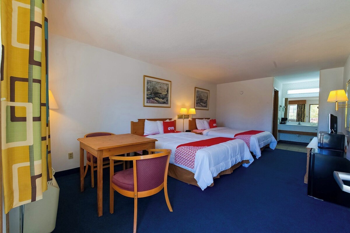 A hotel room with two beds, a wooden table, a chair, a desk with a computer, and a bathroom visible through an open door.