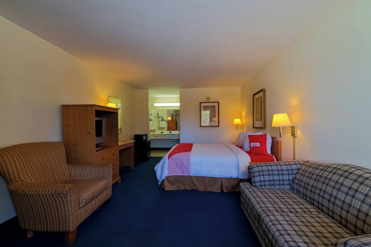 A hotel room with a bed, two armchairs, a wooden cabinet, a desk, a mirror, and a bathroom visible in the background.