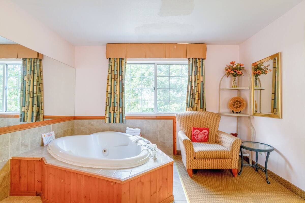 A bathroom with a wooden bathtub, a chair, a small table, and a mirror.