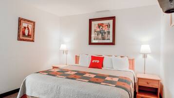 Habitación de hotel con una cama, dos mesitas de noche con lámparas, un almohadón rojo y dos cuadros enmarcados en las paredes.