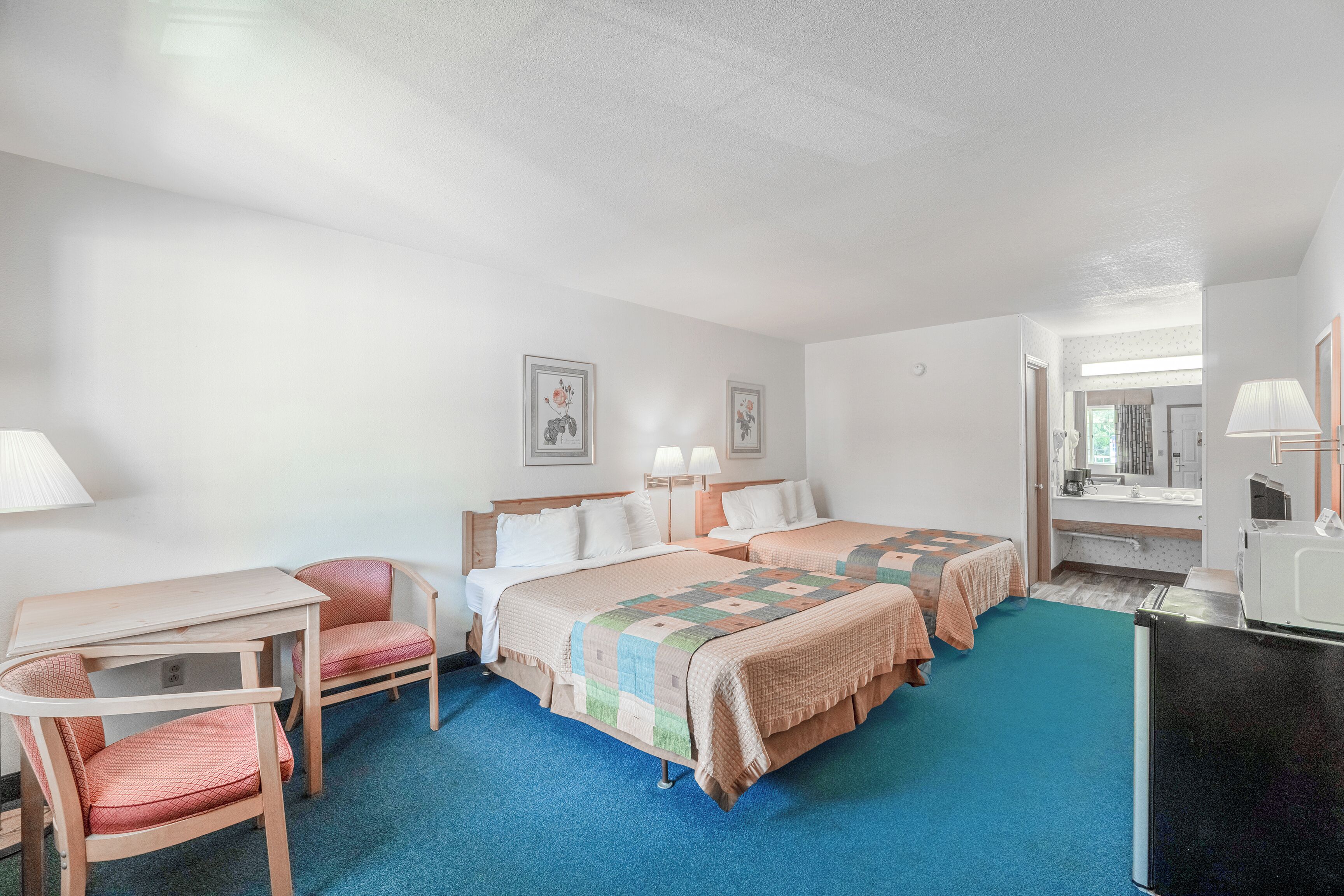A hotel room with two beds, a desk, a chair, a refrigerator, and a bathroom visible through an open door.