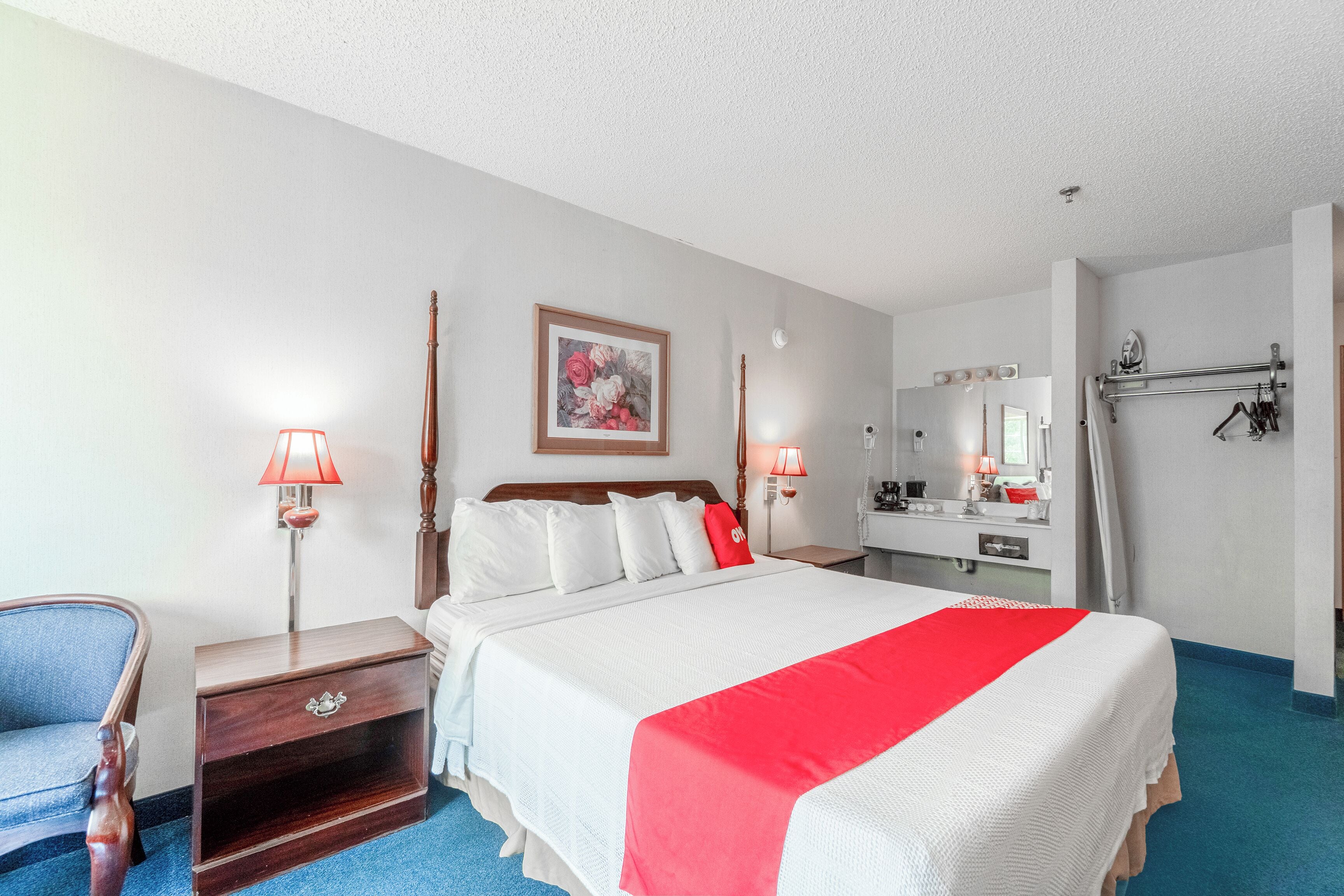 A hotel room with a bed, bedside lamps, a wooden nightstand, a chair, a mirror, and a bathroom visible through an open door.