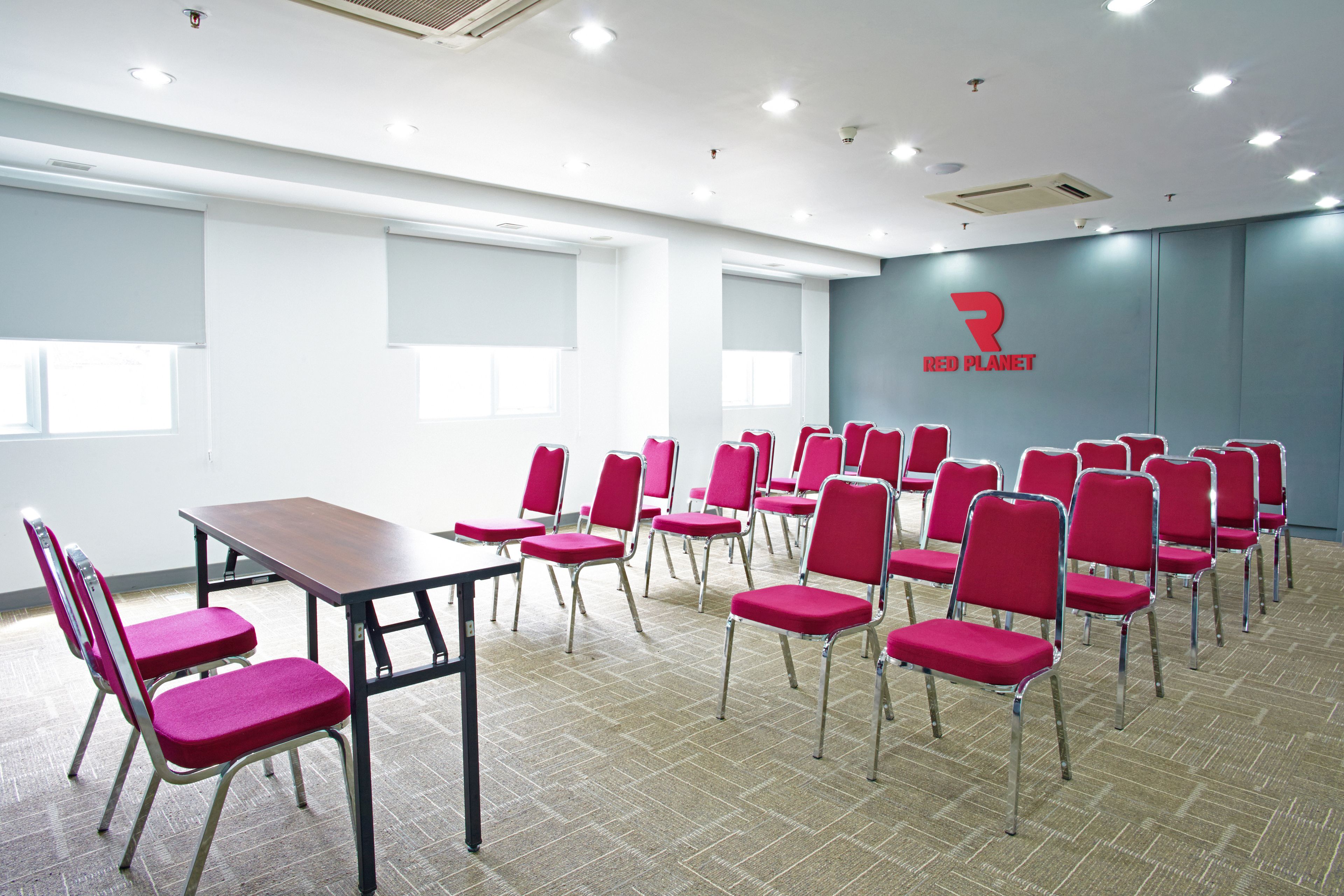 Une salle de conférence avec une longue table et des rangées de chaises rouges, un logo sur le mur et de grandes fenêtres.