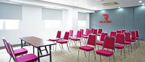 Une salle de conférence avec une longue table et des rangées de chaises rouges, un logo sur le mur et de grandes fenêtres.