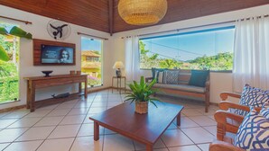Uma sala de estar espaçosa com teto de madeira, luminária trançada grande, televisão, banco de madeira com almofadas e vista para o verde lá fora.