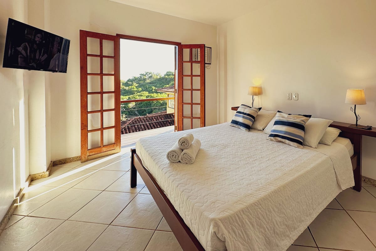 Ein Hotelzimmer mit einem Bett, zwei Nachttischlampen, einem an der Wand montierten Fernseher und einer Glastür zum Balkon.