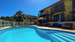 Piscina com azulejos azuis e translúcidos, rodeada por espreguiçadeiras e um edifício com varandas.