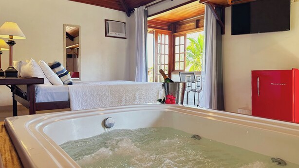 Quarto de hotel com uma banheira grande, uma cama com cobertura branca, um refrigerador vermelho e uma mesa com uma garrafa de champanhe.