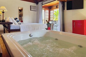 A hotel room with a large bathtub, a bed with a white cover, a red refrigerator, and a table with a bottle of champagne. - Pousada MAR João Fernandes (Búzios)
