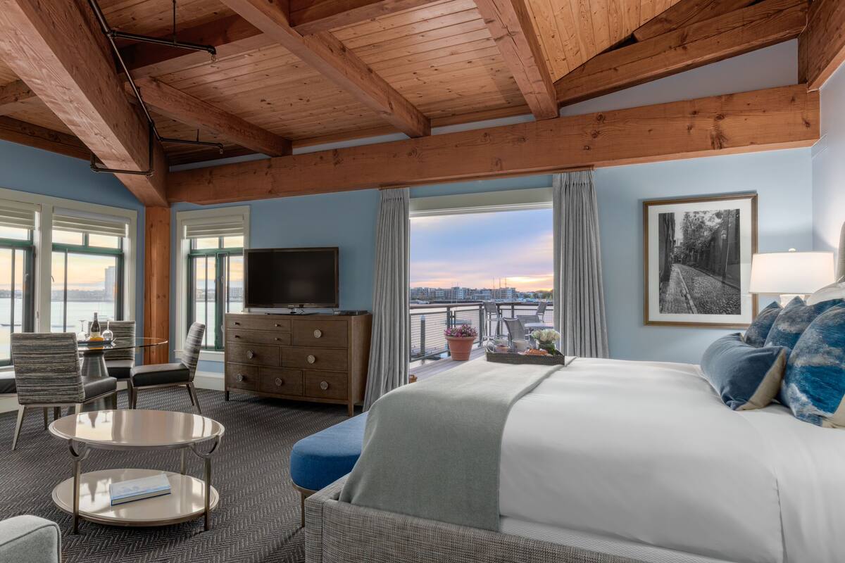 A hotel room with a large bed, a television, a dining area with a table and chairs, and a view of the city through large windows.