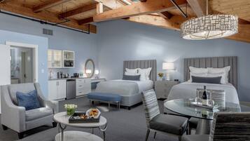 A modern hotel room with a large bed, two bedside tables, a sofa, a small round table, and a kitchenette.