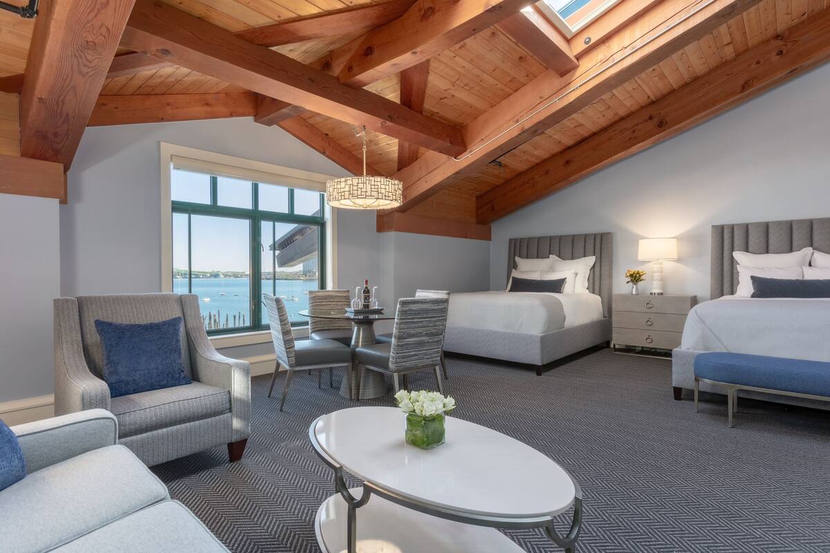 A hotel room with a high ceiling, wooden beams, a large window, a dining area, two beds, and a small table with a vase of flowers.