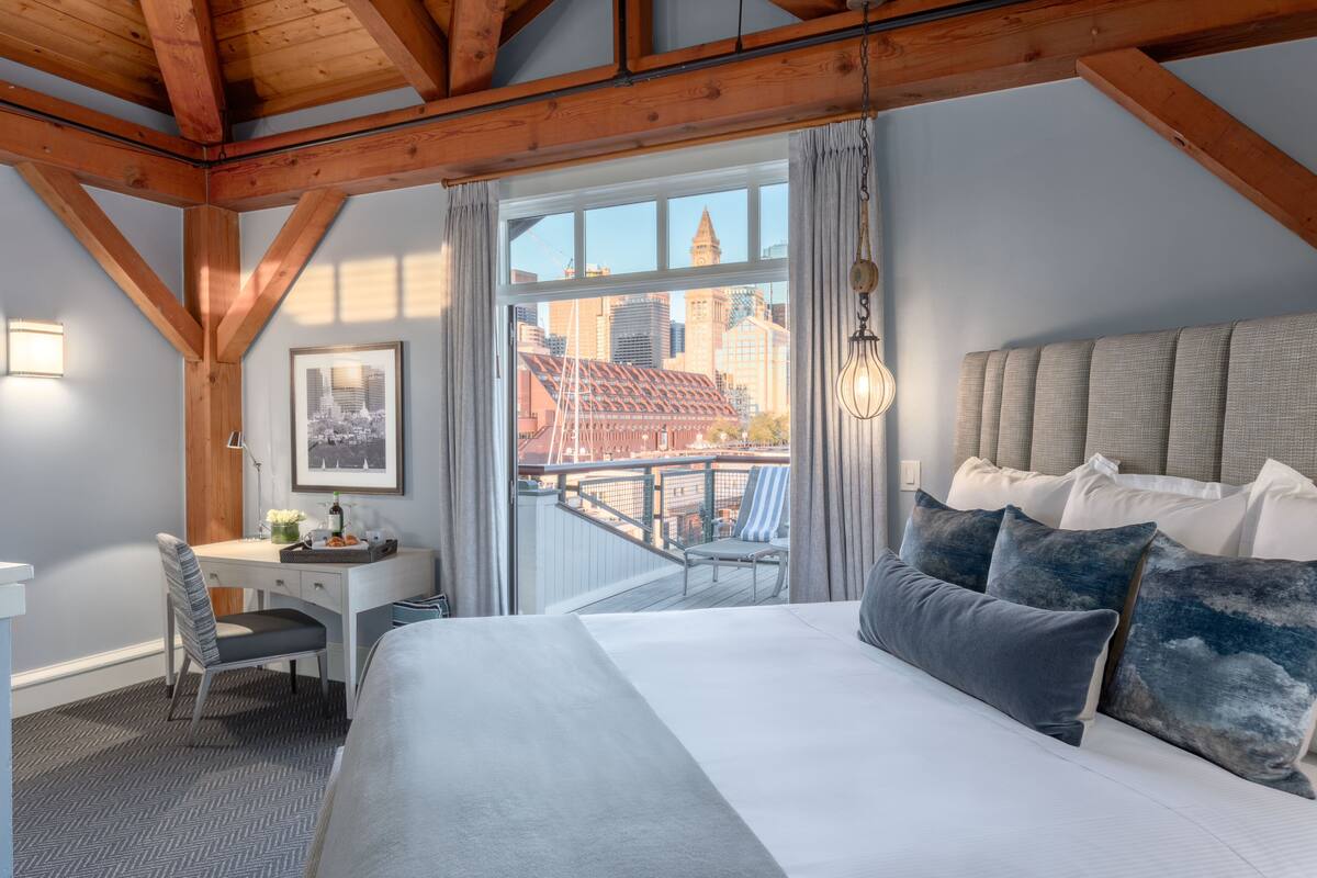 A bedroom with a large bed, a desk, and a chair. It has a view of a cityscape through a large window.