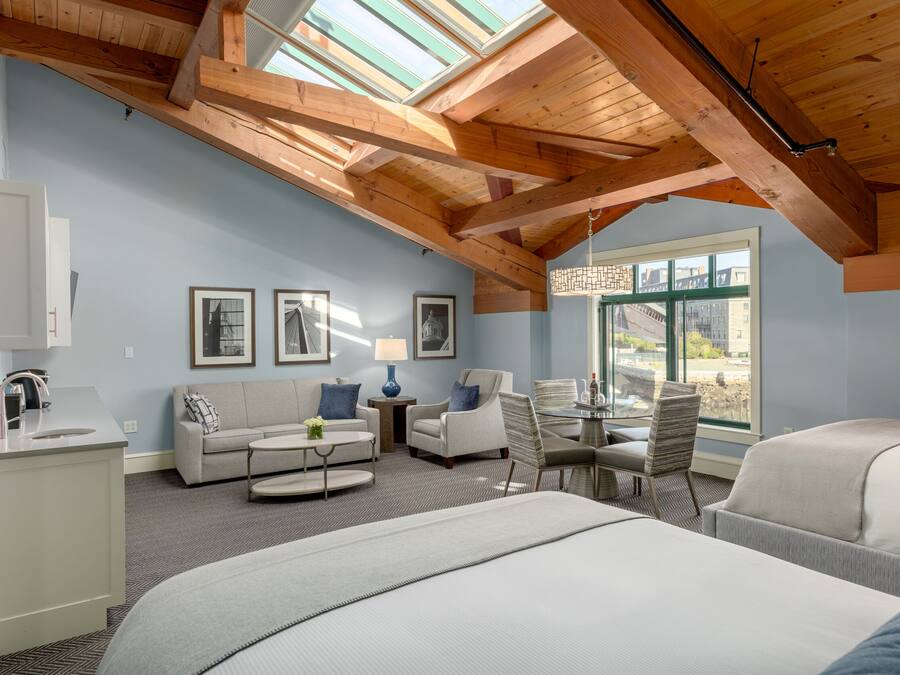 A spacious room with a high ceiling, wooden beams, a large window, a sofa, a dining table, and a bed.