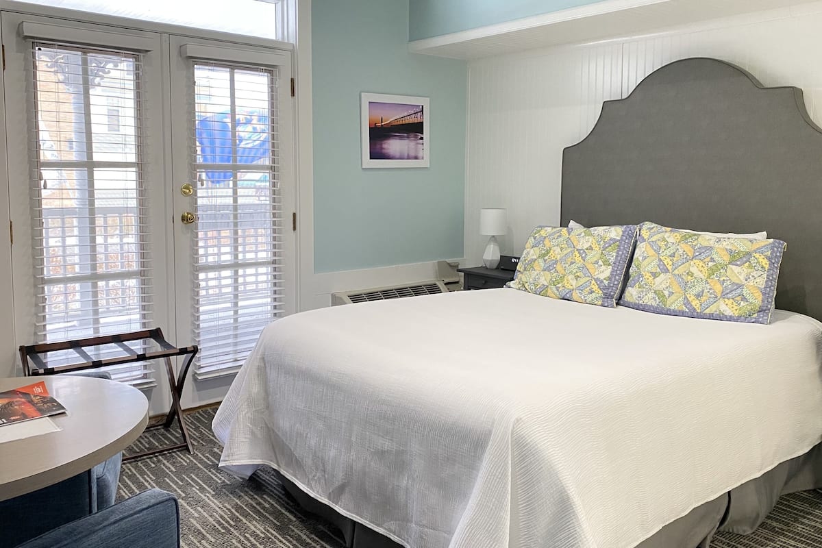 Habitación de hotel con cama, escritorio, silla, ventana con persianas e imagen en la pared.