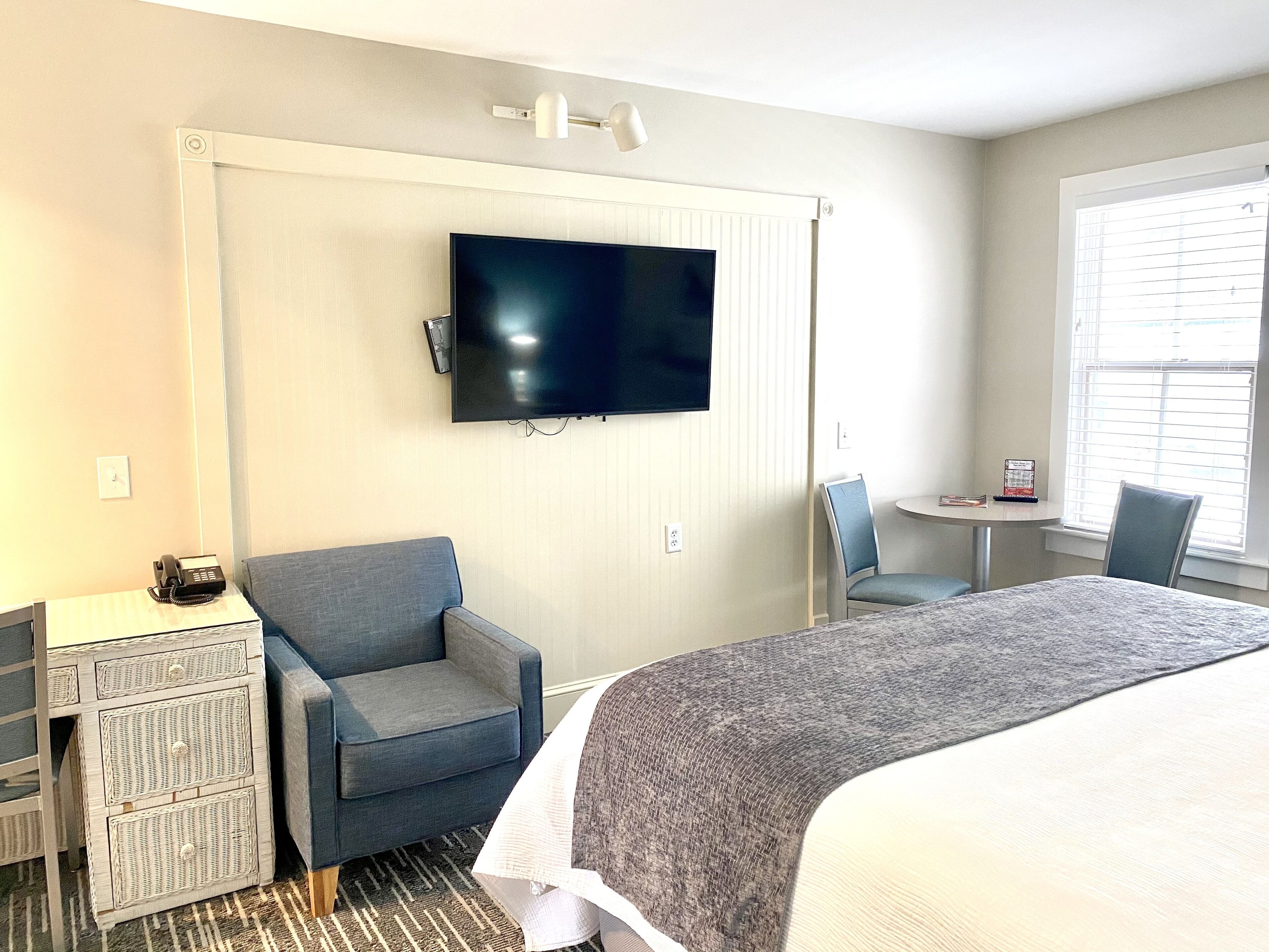 A hotel room with a bed, a desk with a telephone, a chair, a TV mounted on the wall, and a window with blinds.
