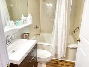 A bathroom with a sink, toilet, and bathtub. There is a shower curtain, a towel rack, and a trash can. - Harbor House Inn (Grand Haven)