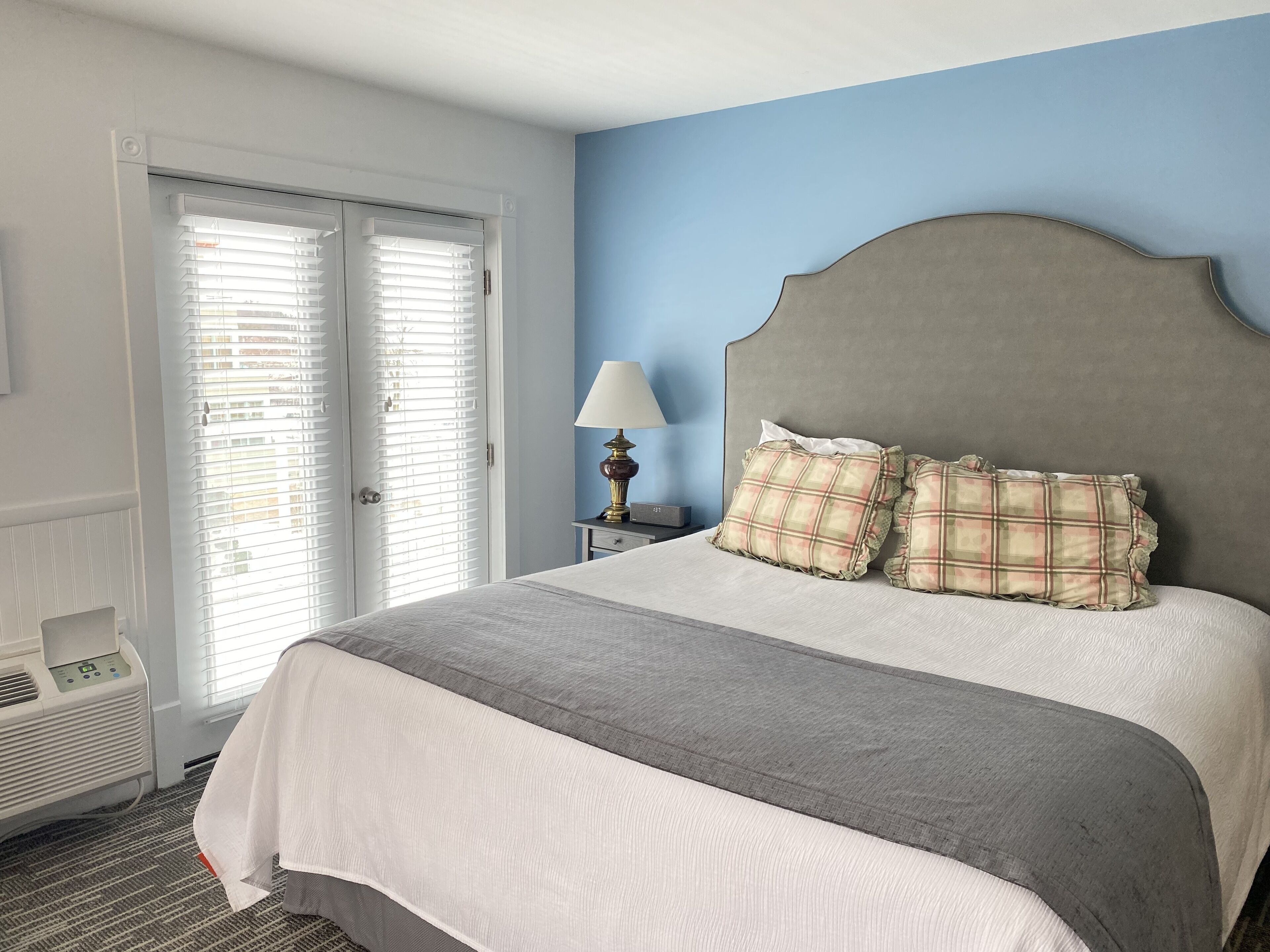 A hotel room with a bed, a bedside table with a lamp, a telephone, and a window with blinds.