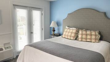 A hotel room with a bed, a bedside table with a lamp, a telephone, and a window with blinds.