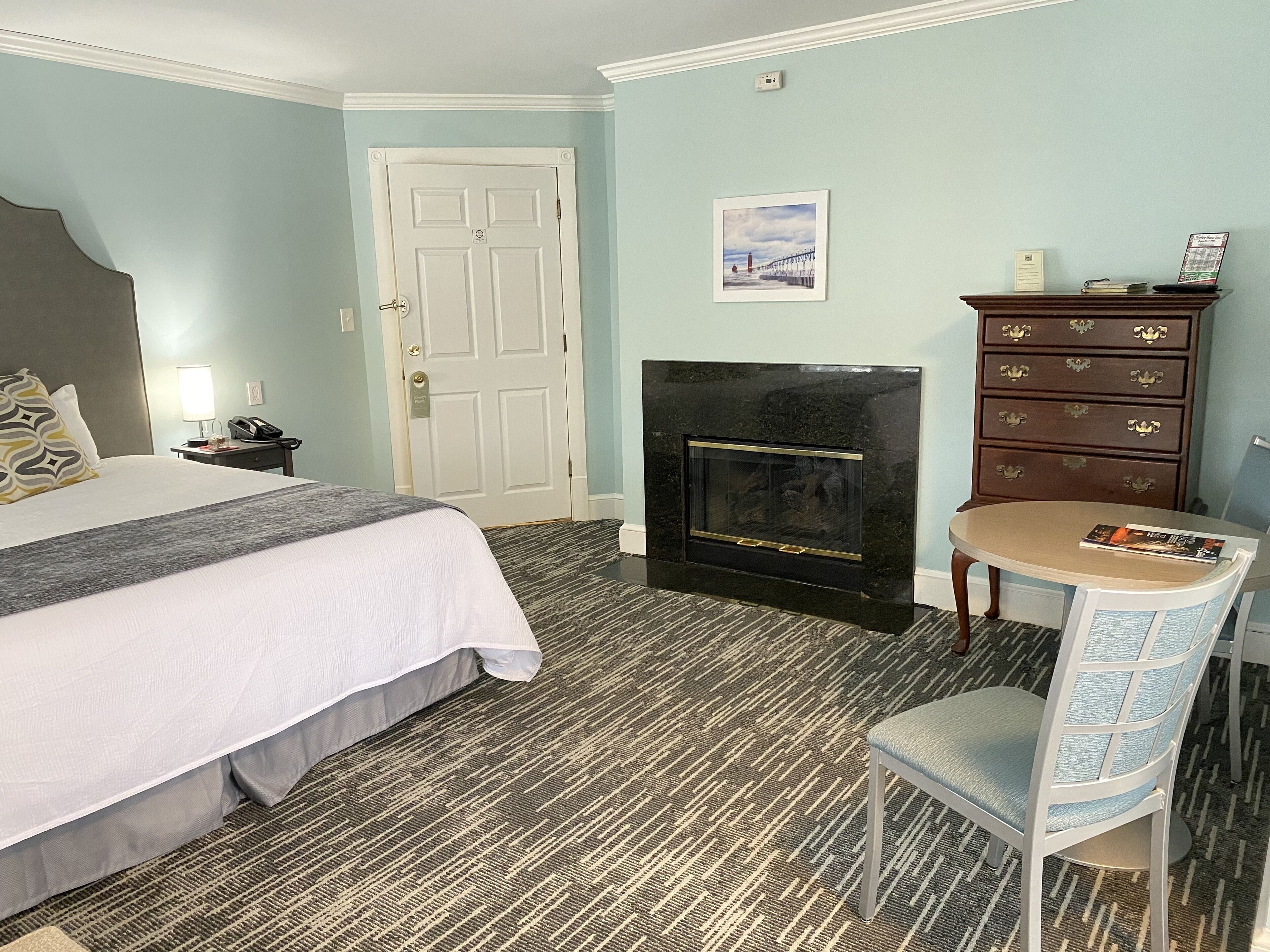 A hotel room with a bed, a desk, a chair, a fireplace, and a dresser.