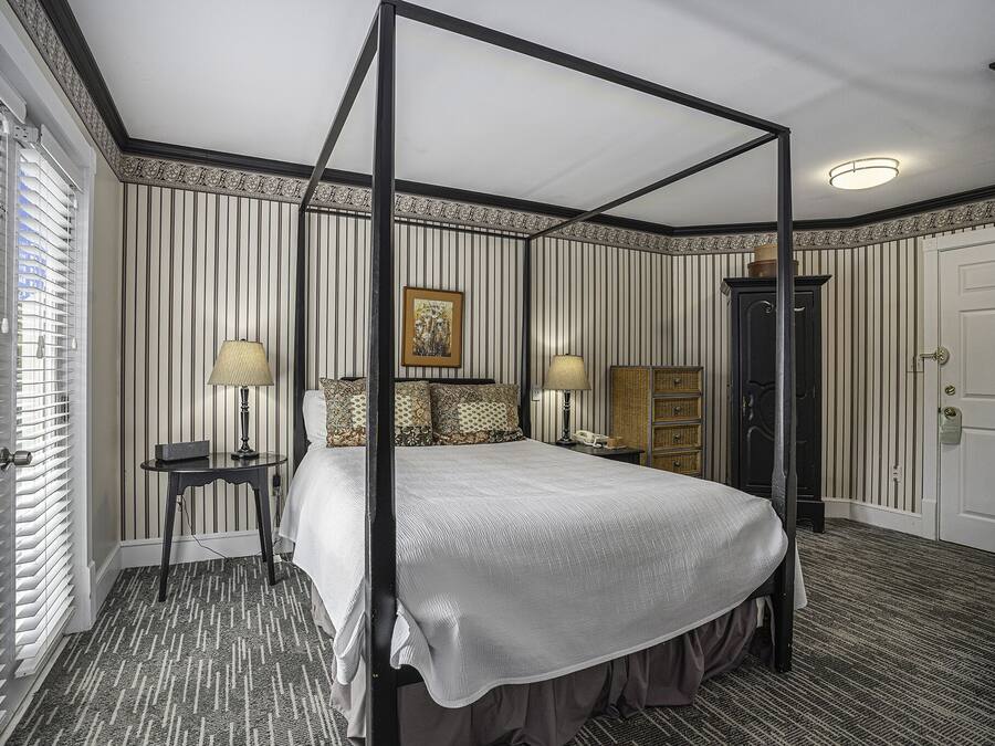 A four-poster bed with a canopy, a bedside table with a lamp, a dresser, and a framed picture on the wall.