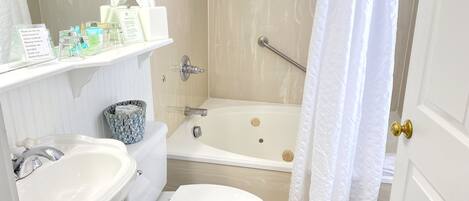 Een badkamer met toilet, wastafel en bad. Toiletartikelen staan op een plankje en het douchegordijn is gedeeltelijk open.