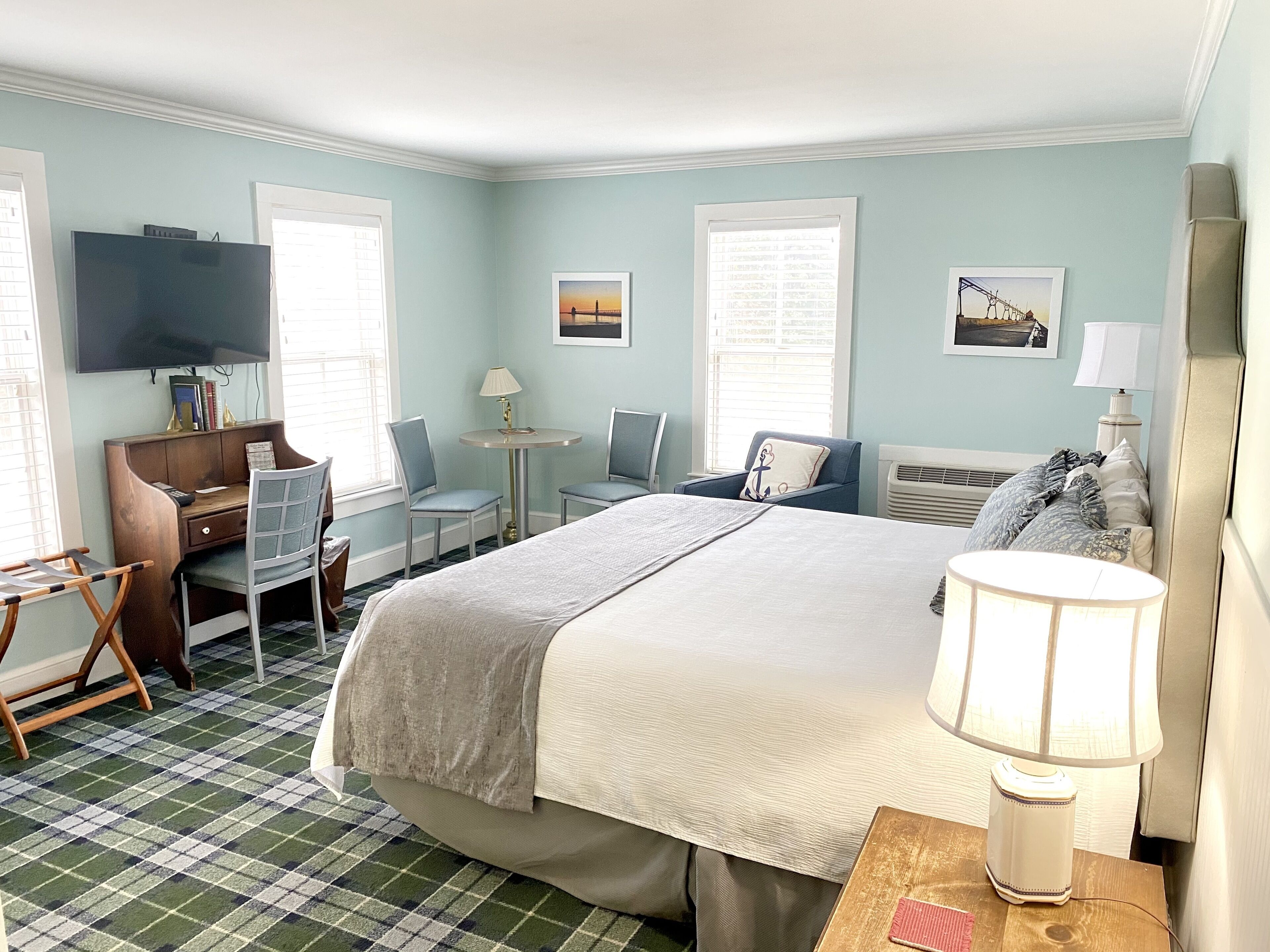 A hotel room with a bed, a desk, a chair, a TV, and two windows.
