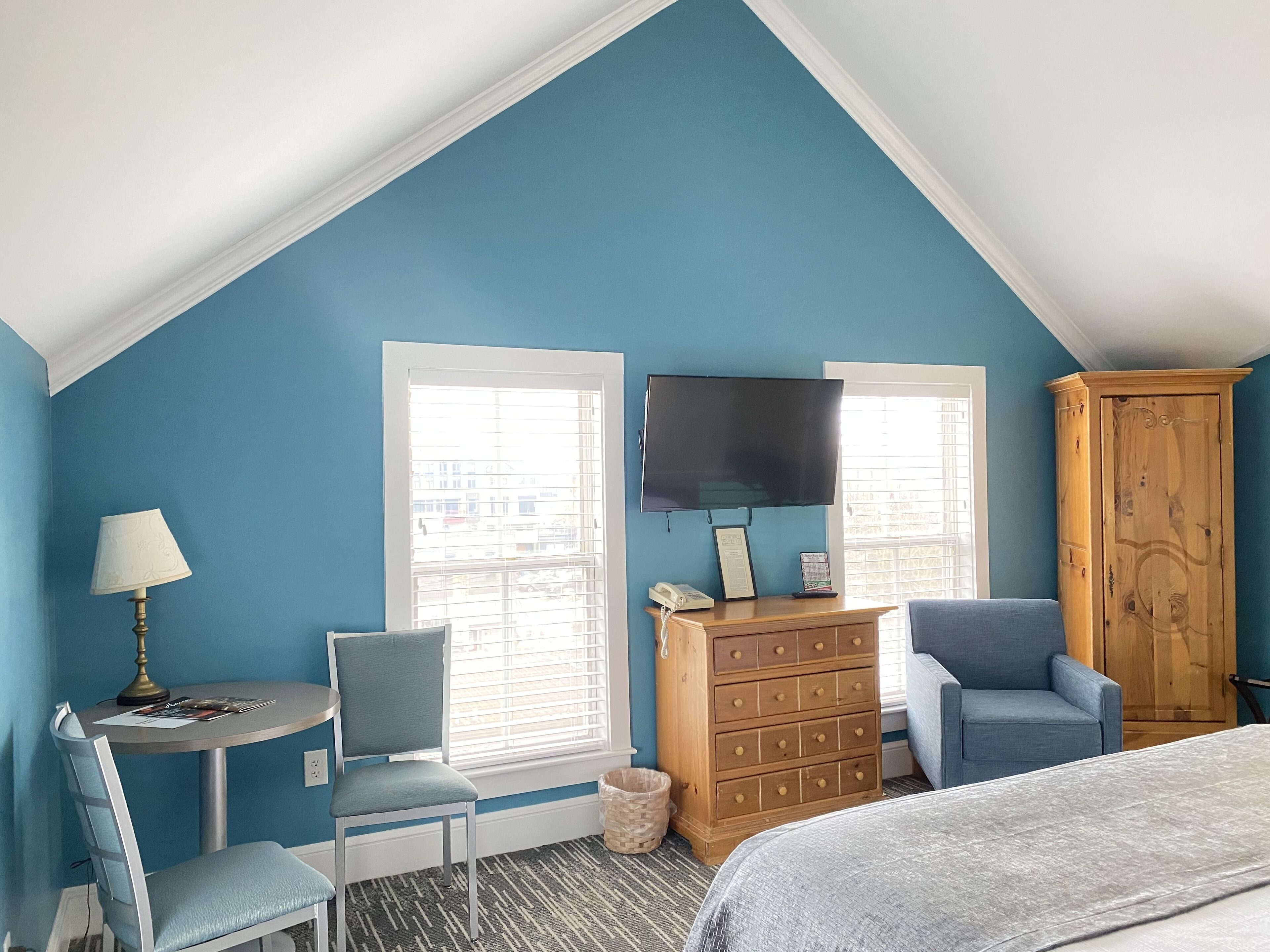 A bedroom with a sloped ceiling, two windows, a television, a wooden dresser, a chair, a desk, and a bed.