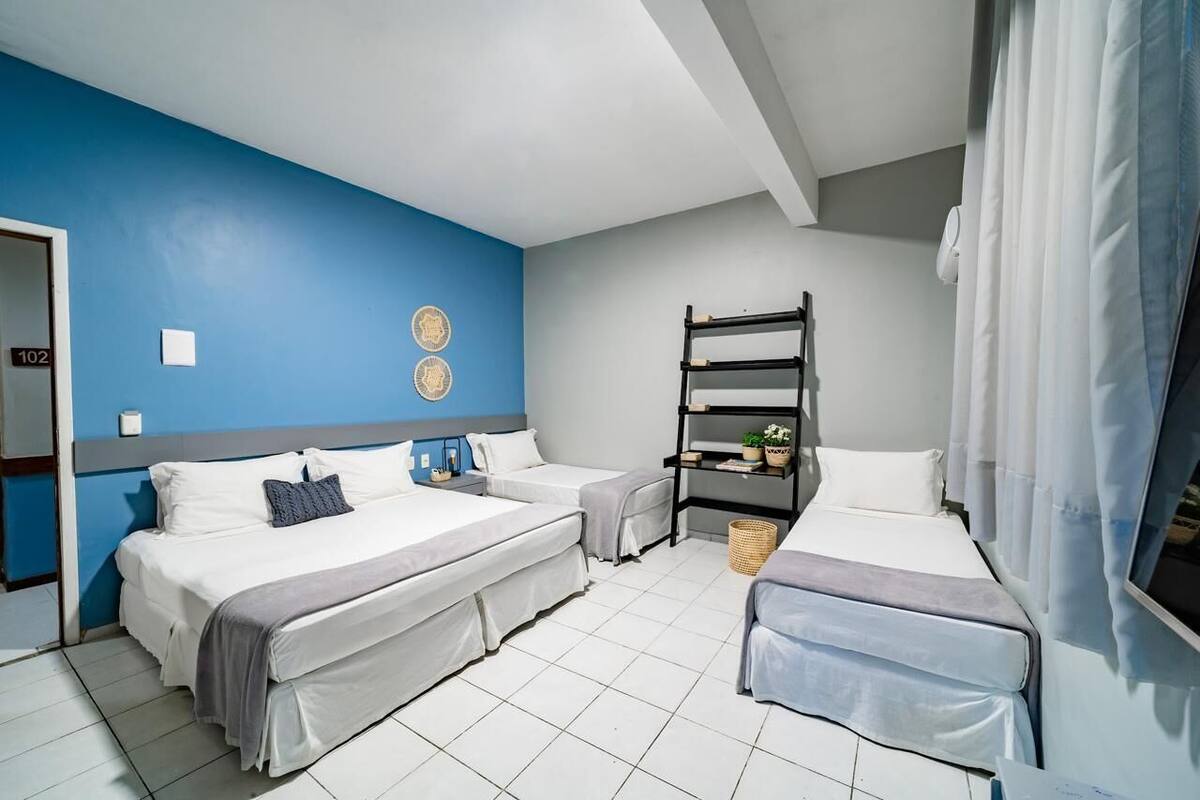 A hotel room with two beds, a bunk bed, a small shelf with a plant, and a window with curtains.
