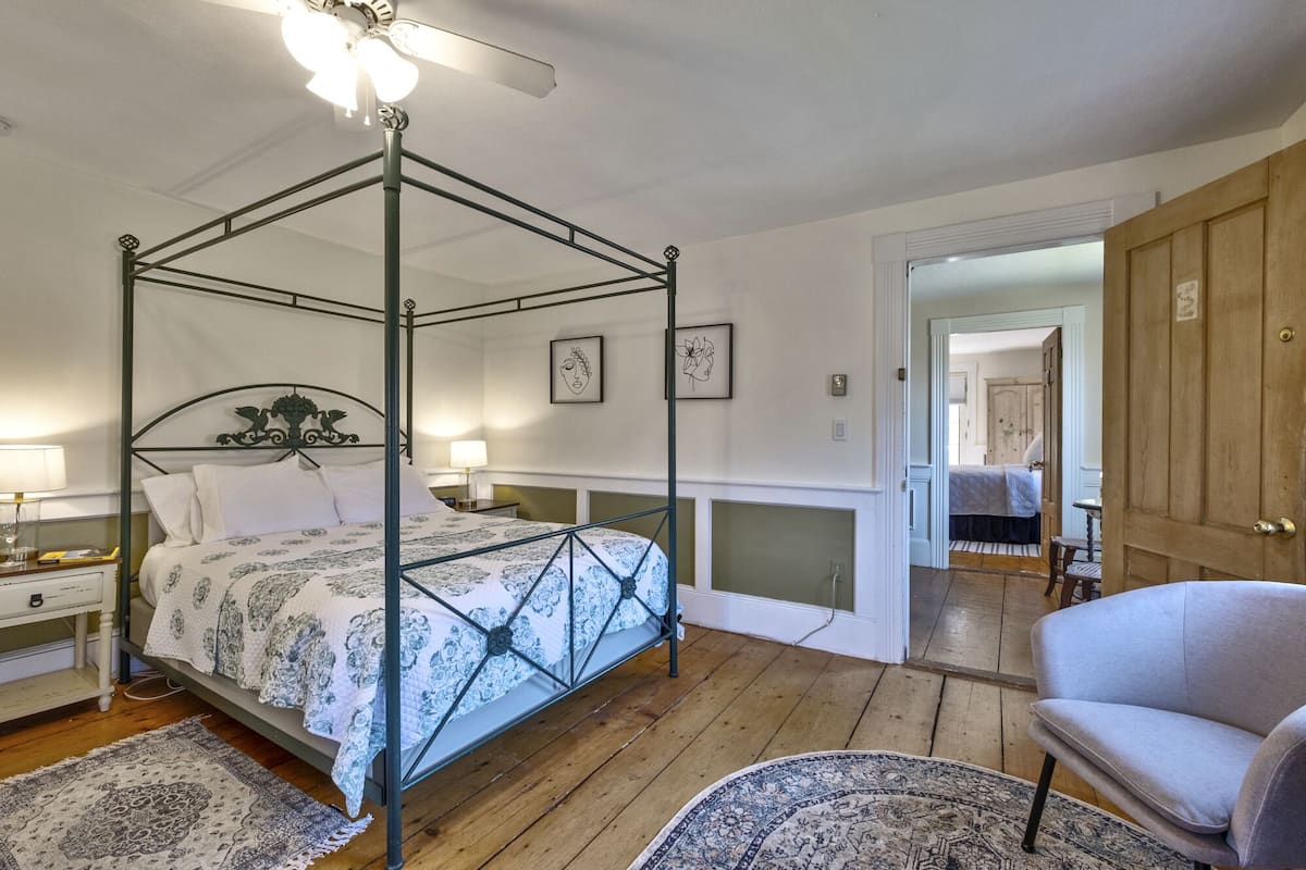 Una cama con dosel con colcha estampada, suelo de madera, una silla y una puerta que da a otra habitación.