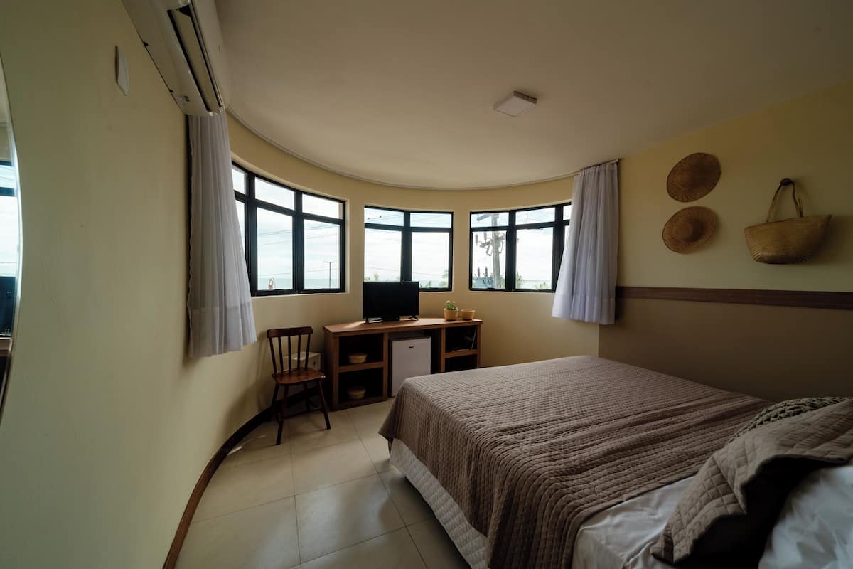 Une chambre à coucher comprenant un lit, un bureau, une chaise et un téléviseur. On y trouve des fenêtres avec des rideaux, un ventilateur de plafond et un panier fixé au mur.