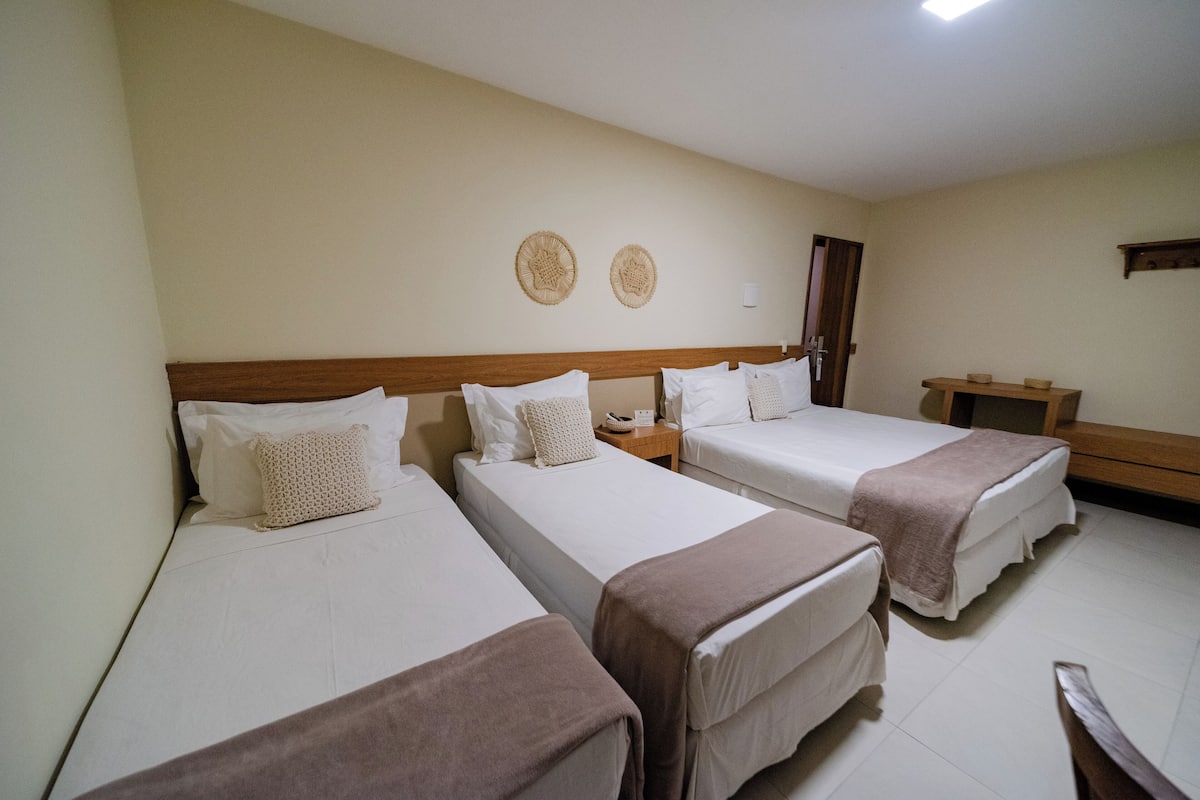Une chambre d’hôtel avec trois lits simples, une tête de lit en bois, une table de chevet et une petite table en bois.