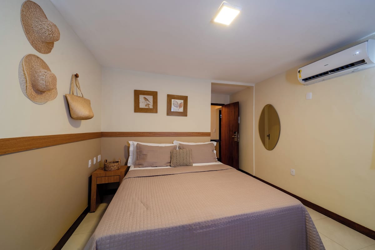 Une chambre à coucher bien rangée, avec un lit, une table de chevet, un climatiseur fixé au mur et deux tableaux encadrés.