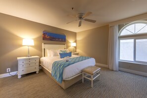 A bedroom with a bed, two bedside tables, a ceiling fan, a window with curtains, and a wall-mounted painting.