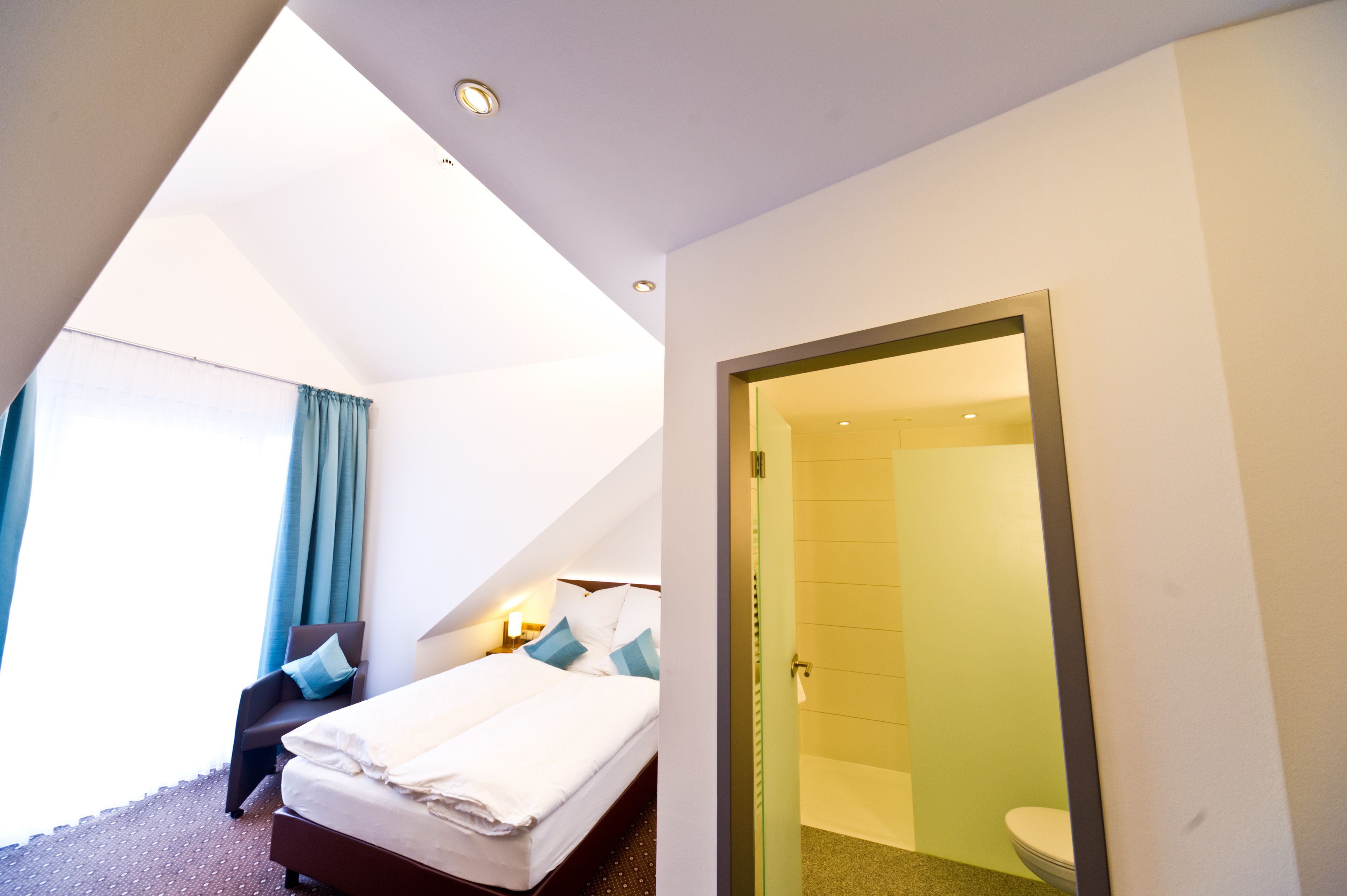 A modern hotel room with a bed, a chair, a desk with a lamp, and a bathroom visible through an open door.