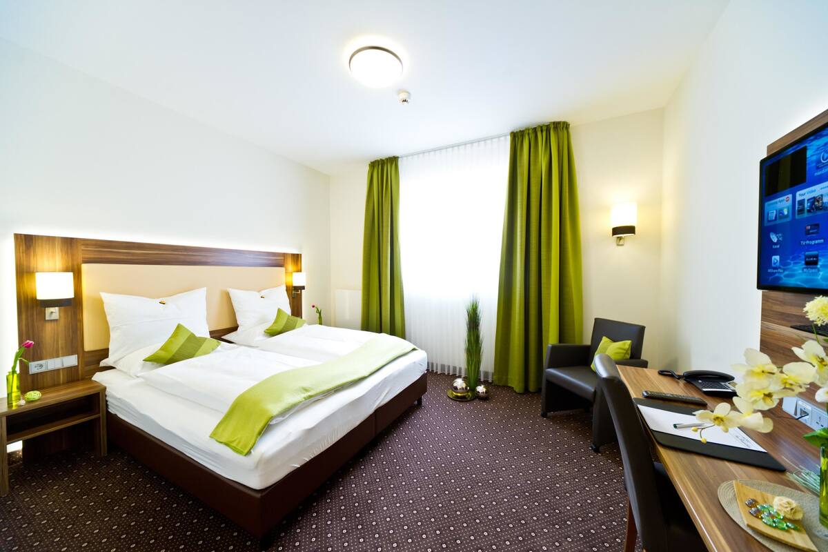 Een hotelkamer met een groot bed, een bureau met stoel, een televisie en een raam met groene gordijnen.