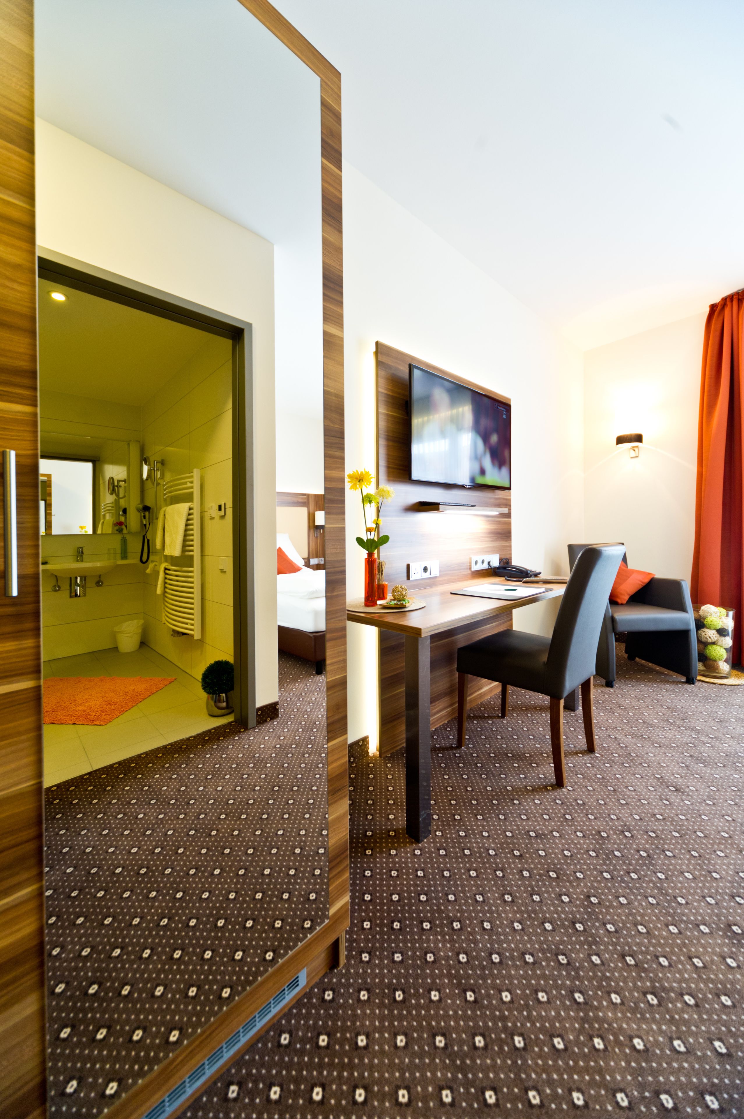 A hotel room with a wooden floor, a desk with a chair, a TV mounted on the wall, a bed with orange pillows, and a bathroom visible through an open door.