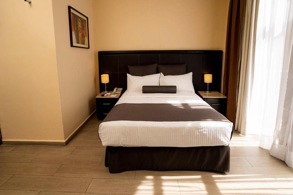 Una camera d'albergo con un grande letto, due comodini, un tavolino e una finestra con tende, oltre a un quadro appeso al muro.
