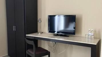 Una habitación pequeña con escritorio, televisión, silla, armario y un cubo de basura.
