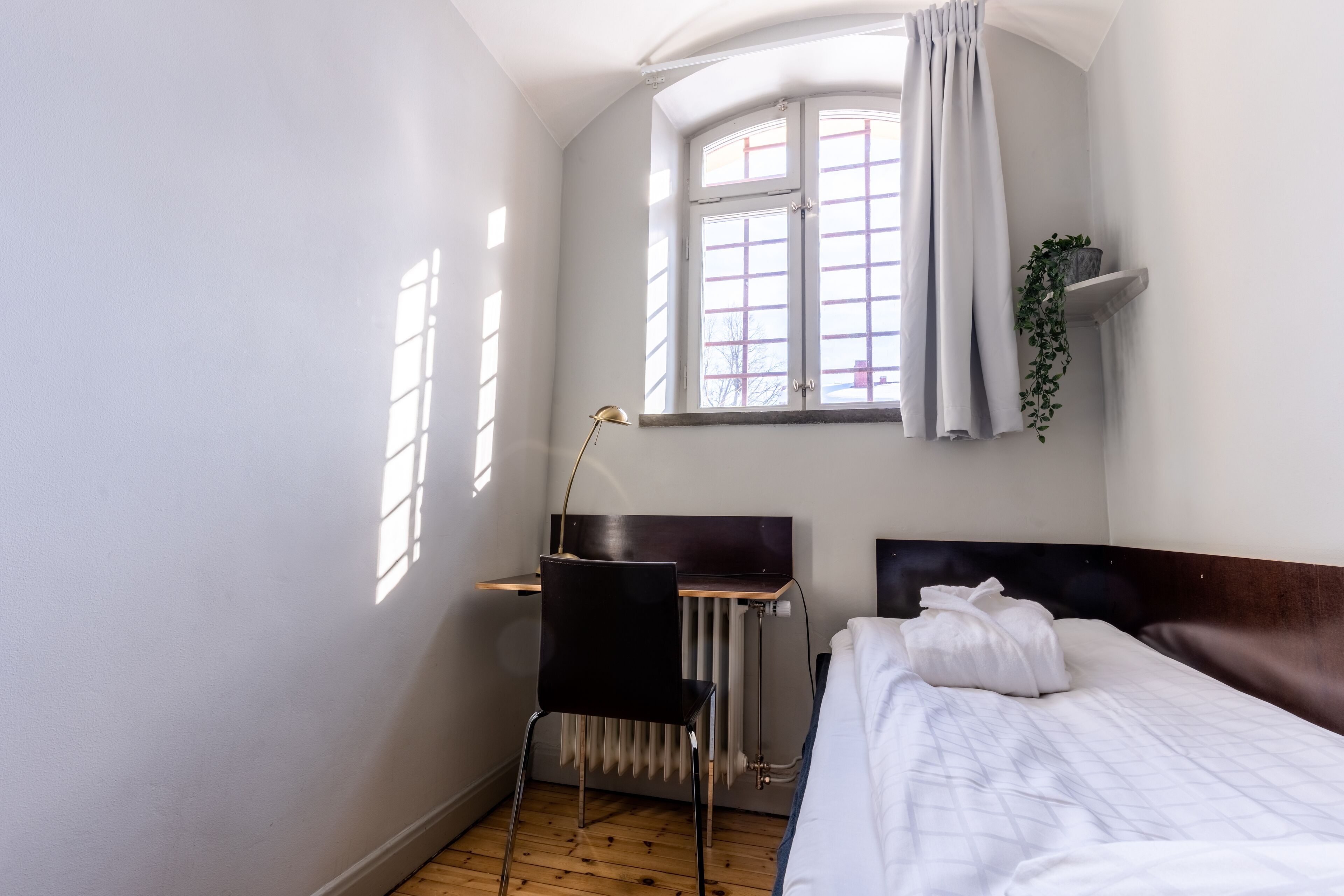 A small, well-lit room with a bed, a desk, and a chair. There is a window with curtains, a radiator, and a plant on a shelf.