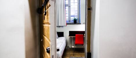 Un pasillo estrecho que conduce a un dormitorio con ventana, un radiador y una silla roja.