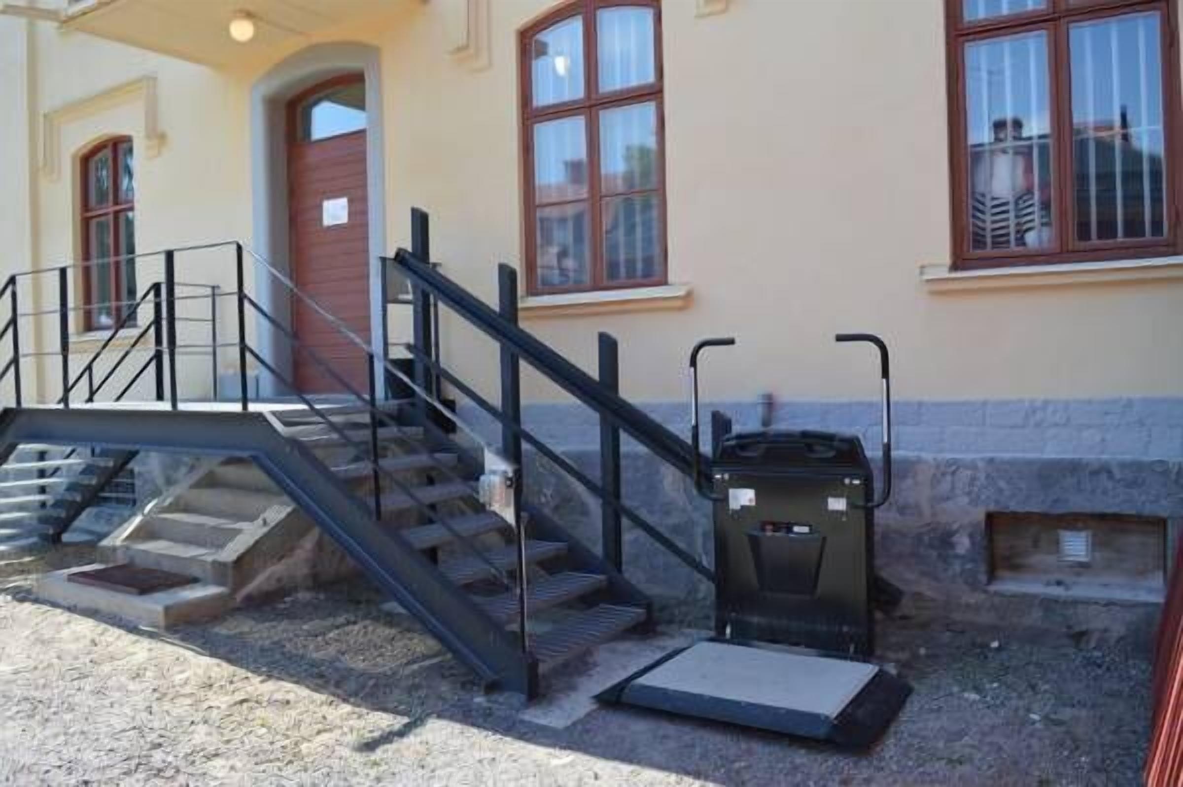 Un edificio con una escalera que lleva a una puerta, un pequeño espacio al aire libre con un contenedor de basura y una ventana con barrotes.