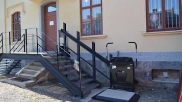 Un edificio con una escalera que lleva a una puerta, un pequeño espacio al aire libre con un contenedor de basura y una ventana con barrotes.
